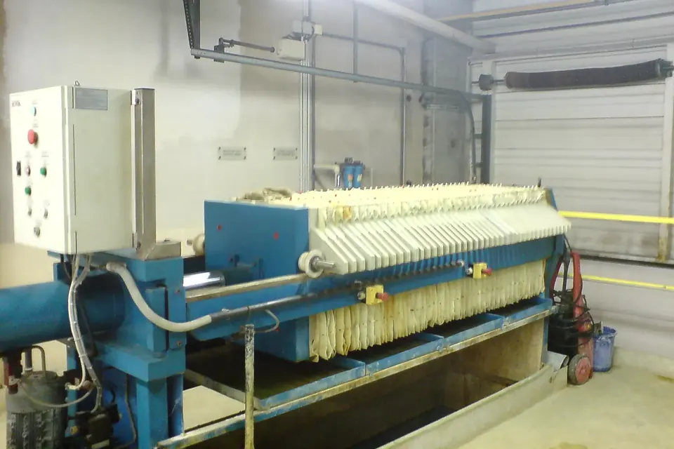 A blue chamber filter press with pale filter plates and a control cabinet in a plant room