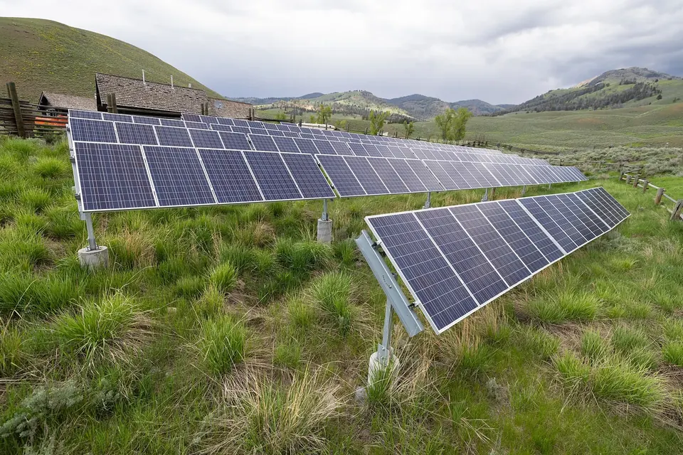 Two rows of ground-mounted solar panels on a grassy slope, with hills and a cloudy sky behind