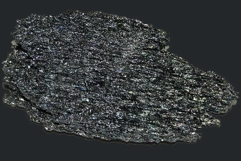 Ein flaches, dunkelgrau glitzerndes Stück kristallines Siliciumcarbid vor graphitgrauem Hintergrund