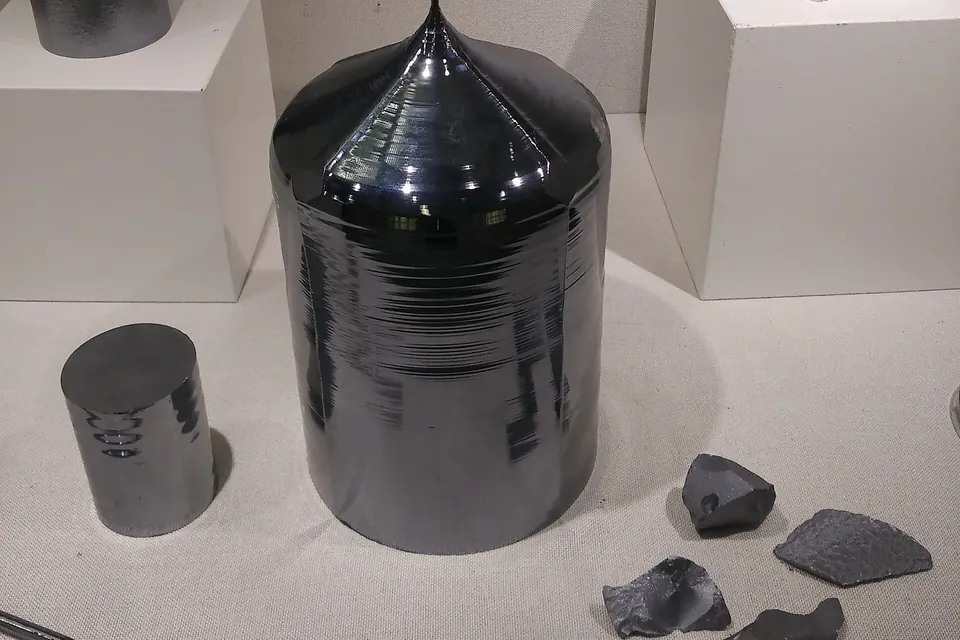 Gezogener monokristalliner Siliciumblock mit kegelförmigem Kopf, daneben ein kurzer Siliciumzylinder und Bruchstücke